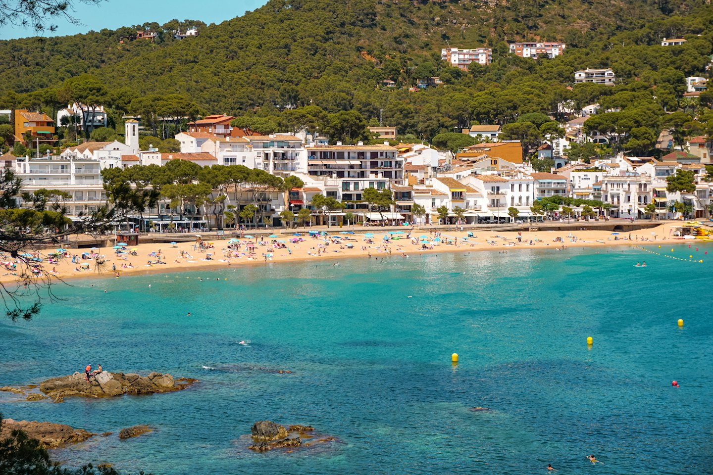 Paese costiero mediterraneo con edifici bianchi, tetti in terracotta e spiaggia affollata.