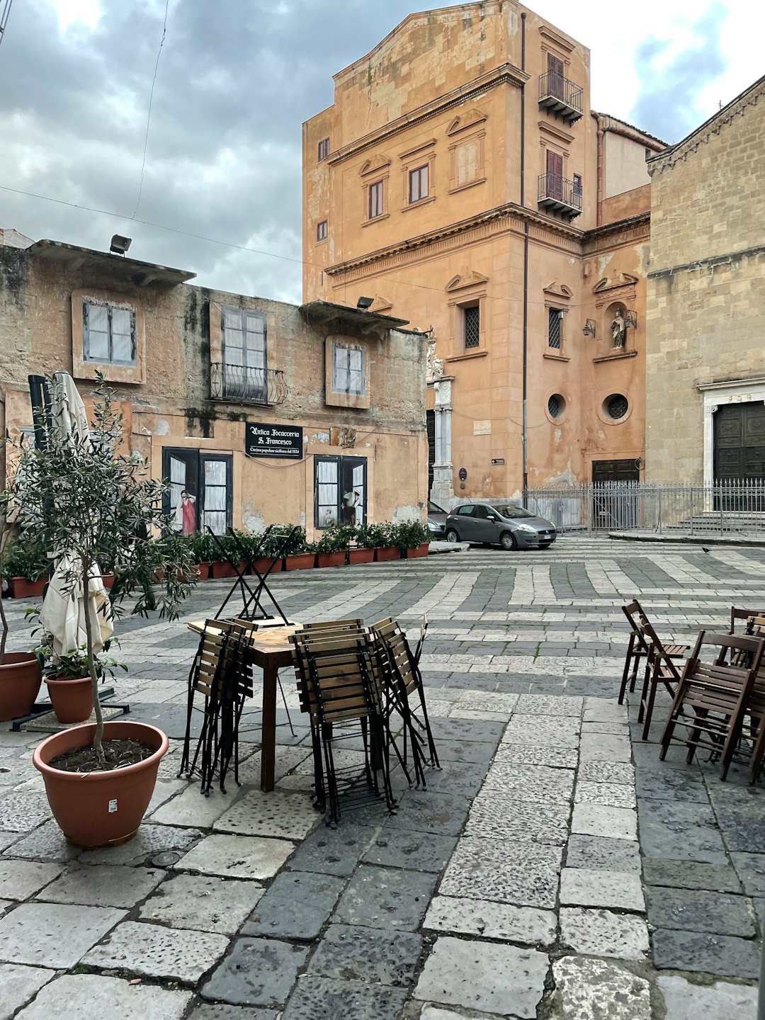 Sedie vuote in un caffè e ulivi in una piazza storica di pietra.